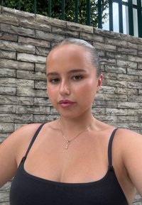 Musta spaghettiolkaiminen toppi, kierrettyä yksityiskohtaa sisältävä kultainen kaulakoru, sileä iho, vaalea tausta, jossa teksturoitu kiviseinä.