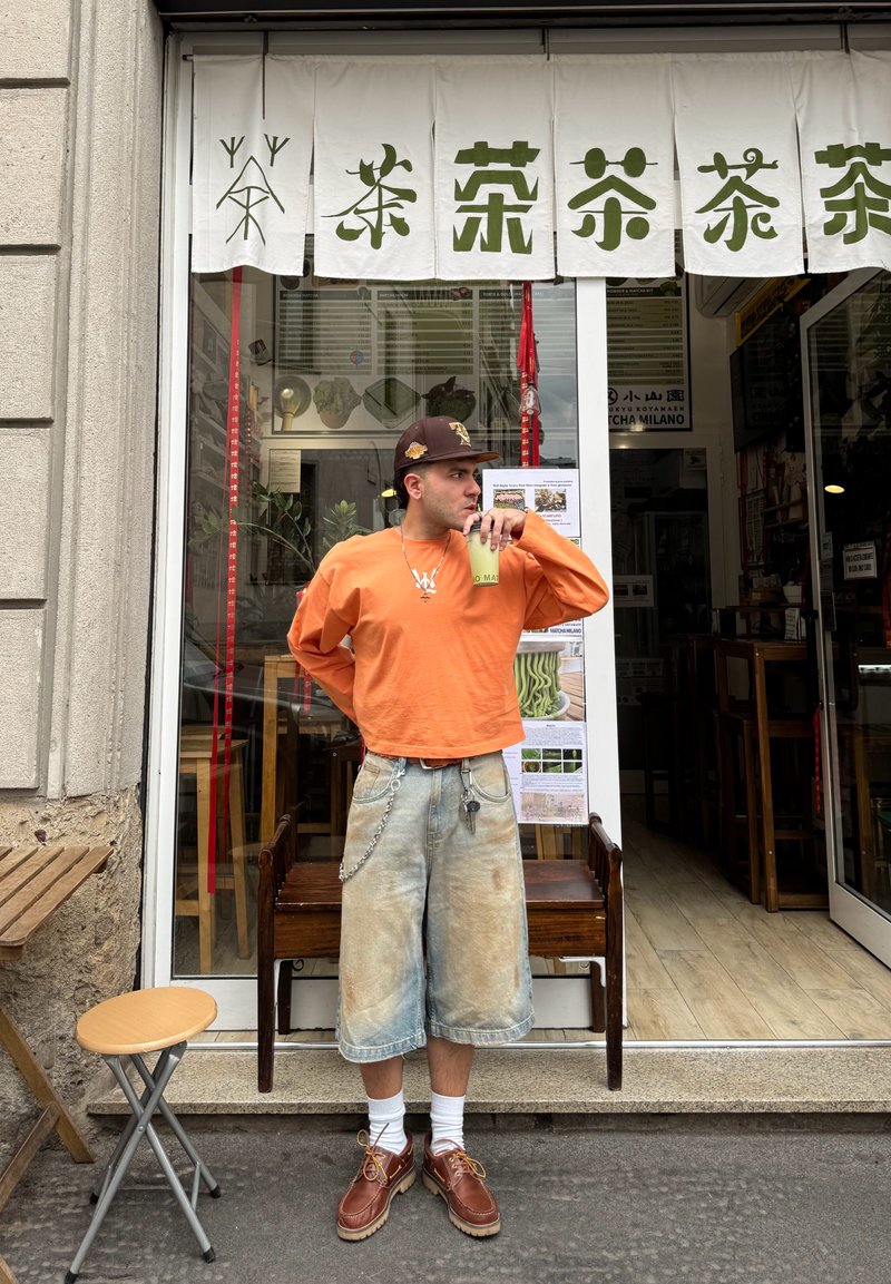 Une personne portant une chemise à manches longues orange vif et un short bleu décoloré se tient devant une boutique de thé, tenant une boisson.