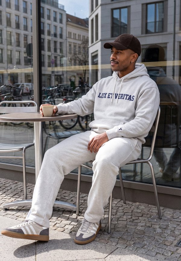 Mand i lysegrå hættetrøje og joggingbukser sidder ved et udendørs cafébord og holder en kaffekop med en bygade, der spejles i vinduet bag ham.