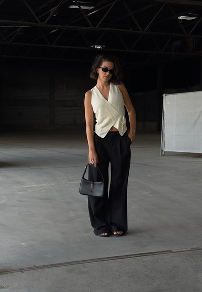 Top blanco con estilo envolvente y un lazo, combinado con pantalones amplios negros. Accesorios incluyen un pequeño bolso negro. Calzado casual visible.
