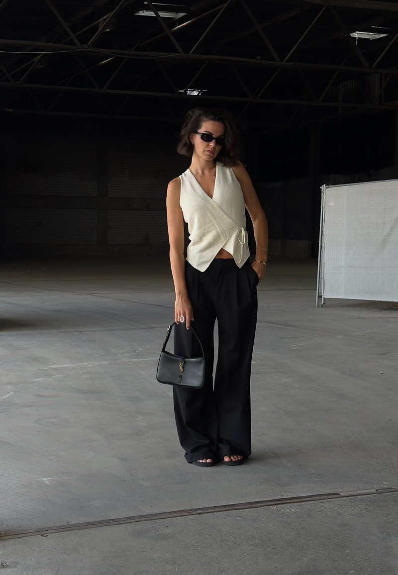 Top bianco a stile avvolto con un nodo, abbinato a pantaloni larghi neri. Accessoriato con una piccola borsa nera. Footwear casual visibile.