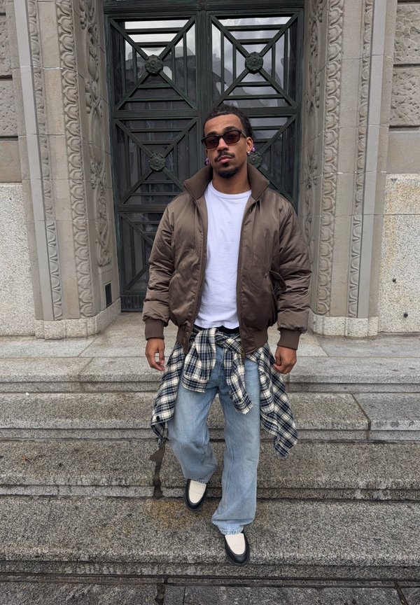 Man wearing sunglasses, brown jacket, white shirt, jeans with a plaid shirt tied around his waist, walking down stone steps in front of an ornate door.