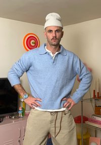 Pull bleu ciel en maille avec un col chemise, pantalon beige à cordon de serrage, et un bonnet blanc. Mains sur les hanches, adoptant une posture neutre.