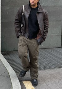 Καφέ δερμάτινο μπουφάν πάνω από ένα μαύρο υφασμάτινο πουλόβερ, συνδυασμένο με χαλαρές ελιές cargo παντελόνες και μαύρα παπούτσια. Το μπουφάν έχει ψηλό γιακά και πλαϊνές τσέπες.