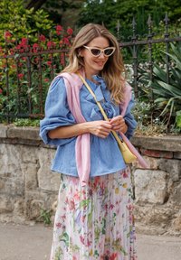 Bluse mit blauen und weißen Streifen und Rüschenkragen, kombiniert mit einem rosa gestrickten Pullover, der über den Schultern drapiert ist, und einem blumenbedruckten, plissierten Rock. Gelbe Umhängetasche.