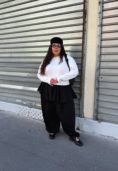 Mujer con cabello largo y rizado, que lleva gafas, sombrero negro, camiseta blanca de manga larga y falda y pantalones negros en capas, de pie junto a persianas metálicas.