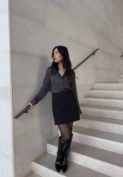 Mujer con cabello largo y oscuro, vestida con una falda negra, camiseta oscura y botas negras, de pie en unas escaleras de mármol, sujetando la barandilla y mirando hacia un lado.