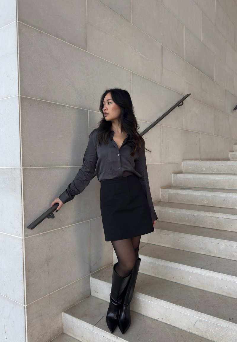 Donna con lunghi capelli scuri, vestita con una gonna nera, una maglia scura e stivali neri, in piedi su una scala di marmo, mentre tiene il corrimano e guarda di lato.