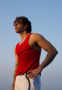 Top de alças riscado vermelho, calças brancas com um cinto texturizado, cabelo cacheado, óculos, em pé contra um fundo de céu limpo.