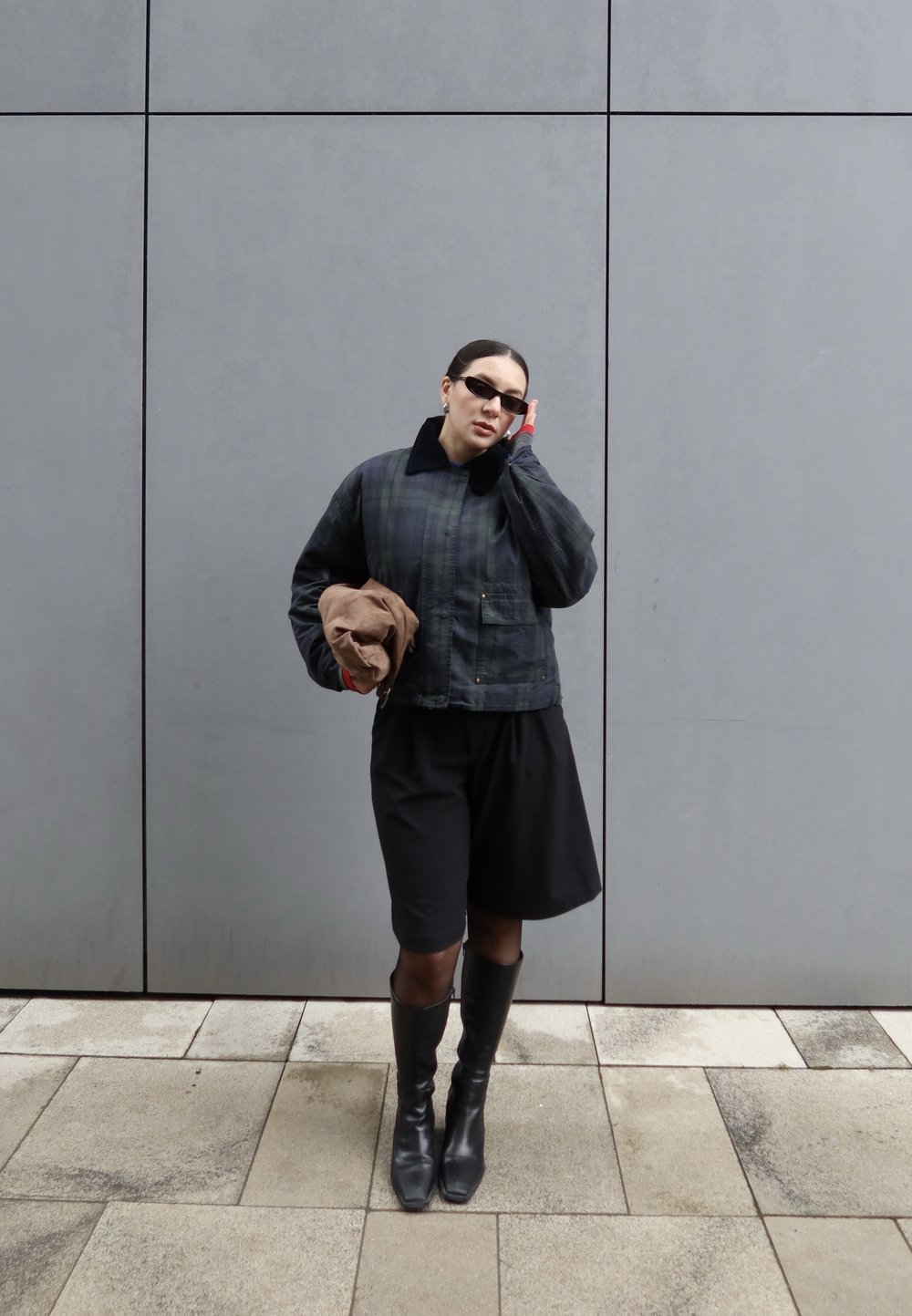 Personne portant une veste à carreaux sombre, un short noir, de grandes bottes noires et des lunettes de soleil, tenant un chapeau marron, debout contre un mur gris.