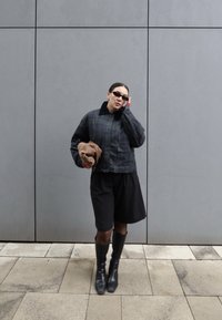 Personne portant une veste à carreaux sombre, un short noir, de grandes bottes noires et des lunettes de soleil, tenant un chapeau marron, debout contre un mur gris.