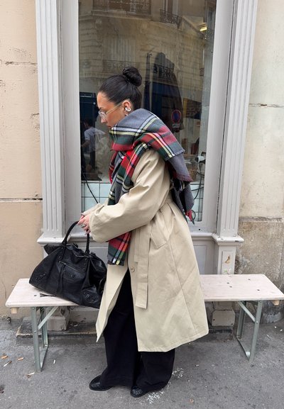 Mujer con abrigo beige y gran bufanda de cuadros roja coloca una bolsa negra en un banco junto a la ventana de un edificio.