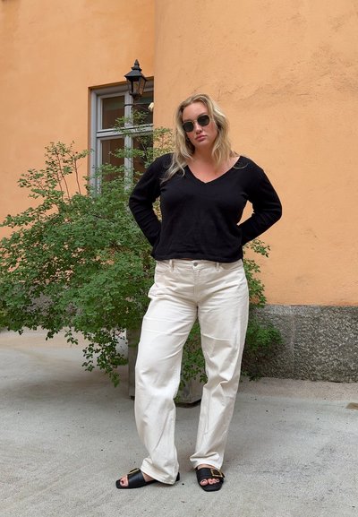 Suéter negro de pico, pantalones crema de pierna ancha y sandalias deslizantes negras. Cabello ondulado; de pie frente a una pared naranja clara y vegetación.