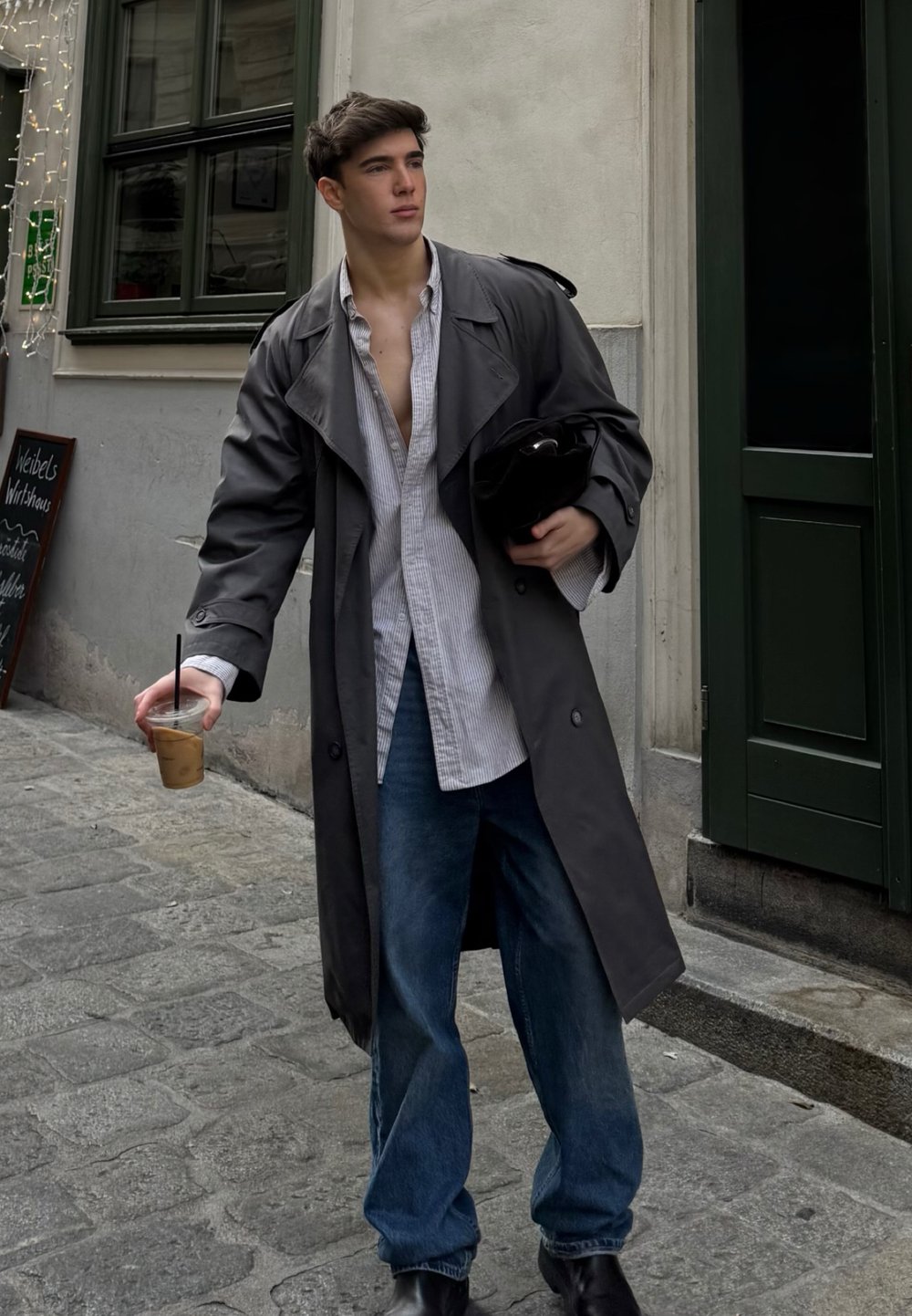 Dunkler, übergroßer Trenchcoat über einem hell gestreiften Hemd, kombiniert mit lockeren blauen Jeans und schwarzen Stiefeletten. Hält ein kaltes Getränk.
