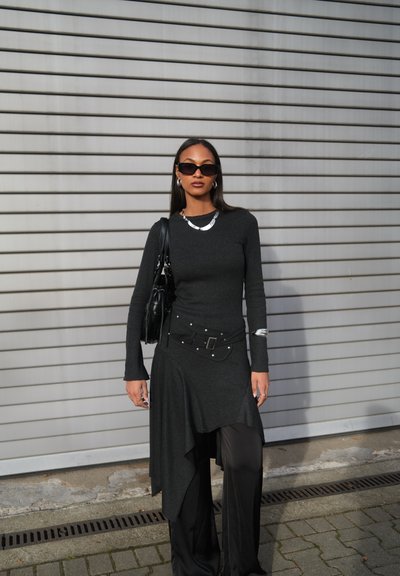 Mujer con vestido asimétrico negro y pantalones anchos, lleva gafas de sol, collar y pulsera plateados, y sostiene un bolso negro, de pie junto a una persiana metálica.