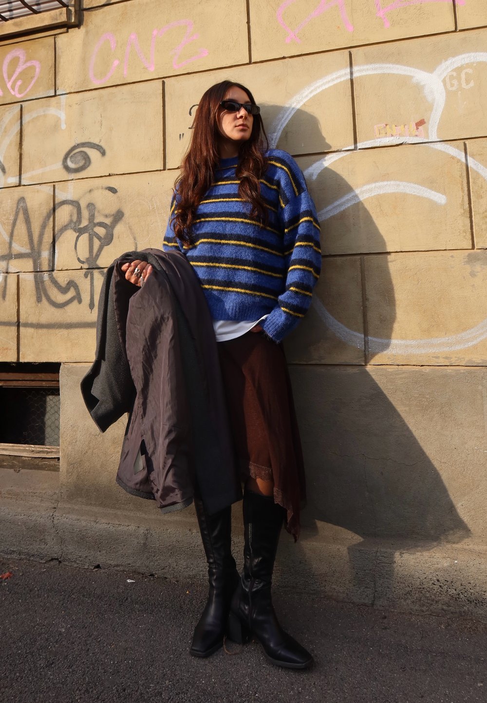 Pull tricot rayé bleu et jaune, jupe marron superposée, manteau gris, et bottes noires à talons hautes jusqu'aux genoux, devant un mur couvert de graffitis.