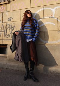 Pull tricot rayé bleu et jaune, jupe marron superposée, manteau gris, et bottes noires à talons hautes jusqu'aux genoux, devant un mur couvert de graffitis.