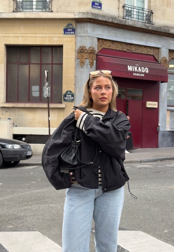 Ung kvinne med blondt hår, svart jakke, stripete skjorte og lyseblå jeans som står i en bygate nær «Mikado Dancing»-stedet.