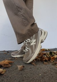 Bež tenisice od mrežastog materijala s kremastim detaljima, gumenim potplatom i vezicama, uparene sa svijetlosmeđim hlačama. Lišće razasuto po tlu.