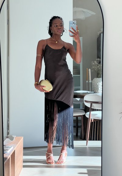 Vestido negro con flecos, diseño sin mangas, textura similar a la seda, vuelo asimétrico, combinado con un clutch dorado y sandalias transparentes con tiras.