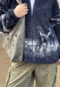 Veste en polaire marine avec motif de montagne blanc, sac en cuir gris avec découpes circulaires, pantalon cargo olive, et plusieurs bagues à la main.