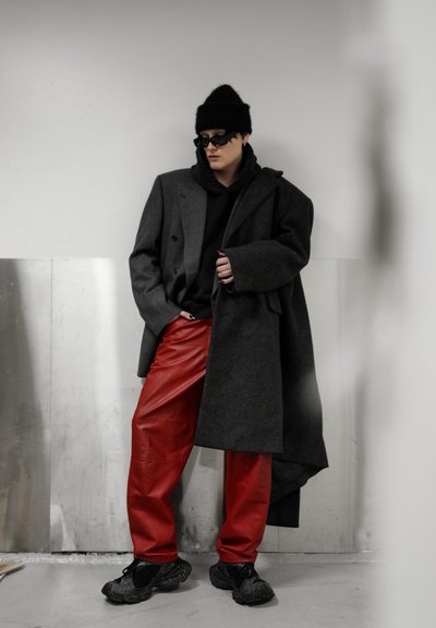 Persona con gorro negro, gafas de sol, abrigos grandes grises y negros, sudadera con capucha negra, pantalones de cuero rojos y zapatillas negras, de pie en interiores.