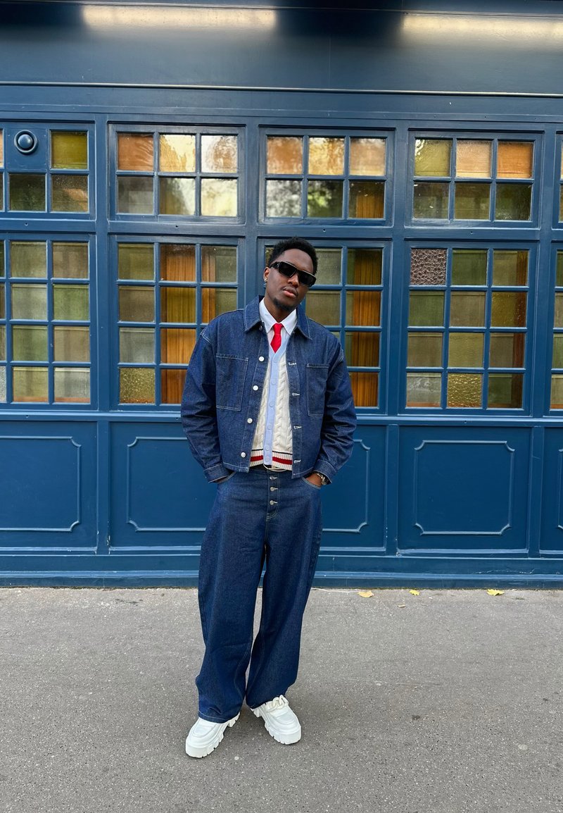Denim jacket and wide-leg jeans in dark blue, layered over a textured white sweater with a red and blue stripe, paired with white shoes.