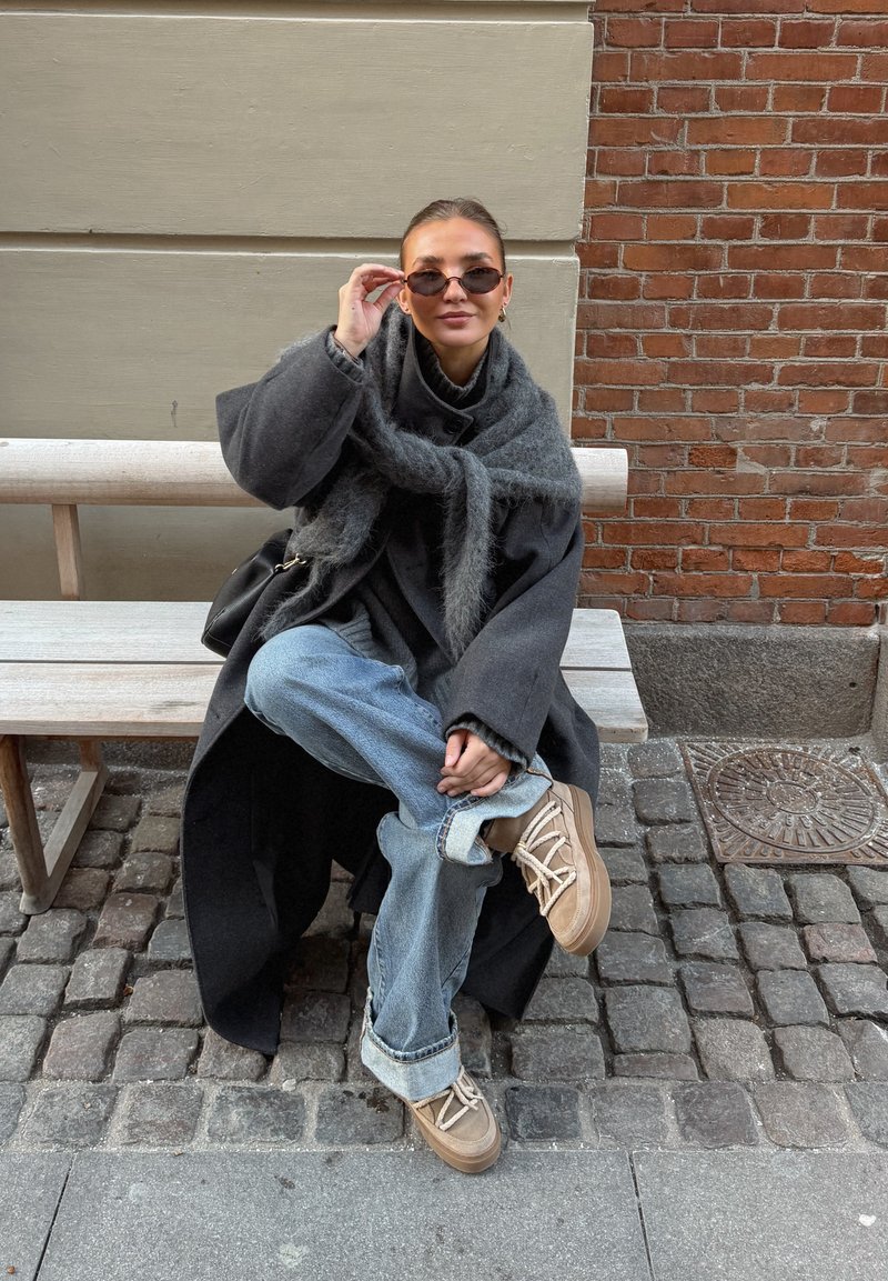 Femme portant un manteau sombre surdimensionné, une écharpe grise, des lunettes de soleil et un jean bleu clair, assise en tailleur sur un banc en bois dans une rue pavée.