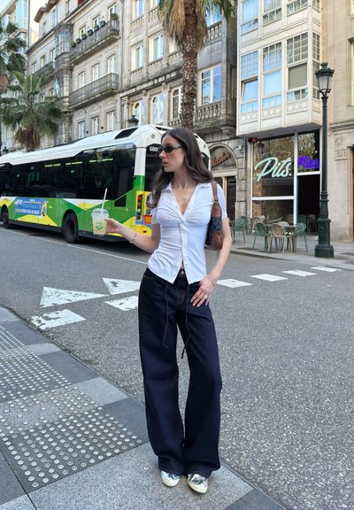 Camisa blanca de botones, jeans oscuros de pierna ancha y zapatos plateados. Sosteniendo una bebida, en una calle de la ciudad con palmeras y un autobús.