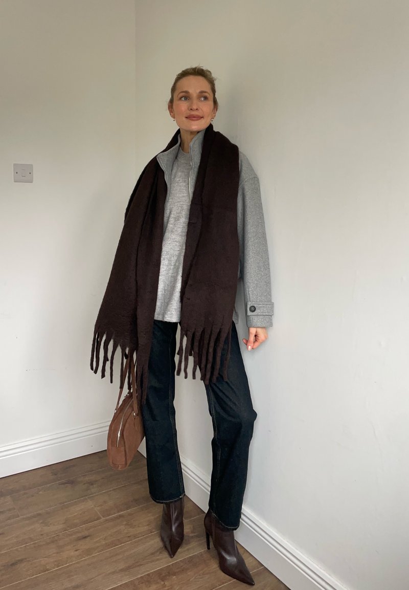 Femme portant un pull gris, un jean foncé, des bottes à bouts pointus marron, une grande écharpe marron foncé, tenant un petit sac à main marron, debout contre un mur blanc.