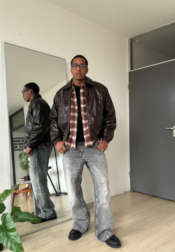 Muž s brýlemi, v hnědé kožené bundě, kostkované košili a šedých džínách stojí uvnitř poblíž zrcadla na celé postavy a zavřených šedých dveří.