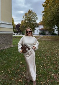 Pull léger crème surdimensionné avec des poignets côtelés, associé à une jupe maxi satin beige clair, un sac à main marron avec un accent pompon et des lunettes de soleil foncées.