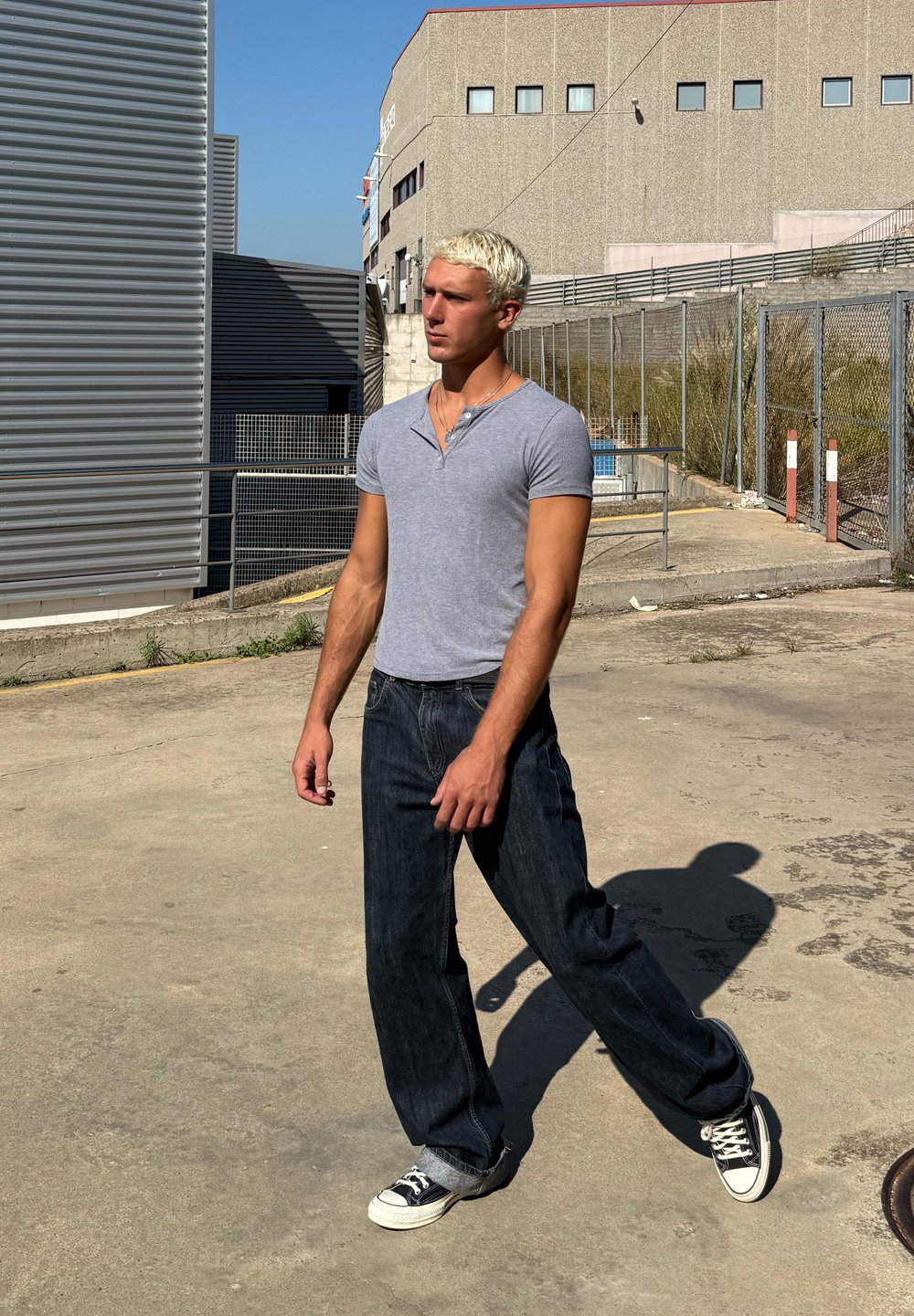 Grå tætsiddende T-shirt, mørkeblå løse jeans og sort-hvide sneakers. Udendørs miljø med en betongulv og industribaggrund.