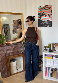 Femme en débardeur sombre et jean large avec des lunettes de soleil, debout près d'une cheminée en marbre rouge, d'un miroir et d'une étagère avec des livres et des appareils photo.