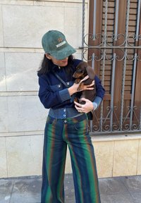 Μαρινισιέ καλυμμένο σακάκι με δύο μπροστινές τσέπες, παντελόνι τύπου wide-leg σε πράσινες και μπλε ρίγες, και πράσινο καπέλο με το λογότυπο "MISSONI". Κρατάει ένα καφέ κουτάβι.