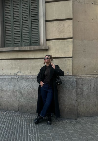 Abrigo largo negro sobre una blusa marrón texturizada, jeans azules y zapatos negros. Los accesorios incluyen gafas de sol y un pequeño bolso negro. Muro de piedra de fondo.