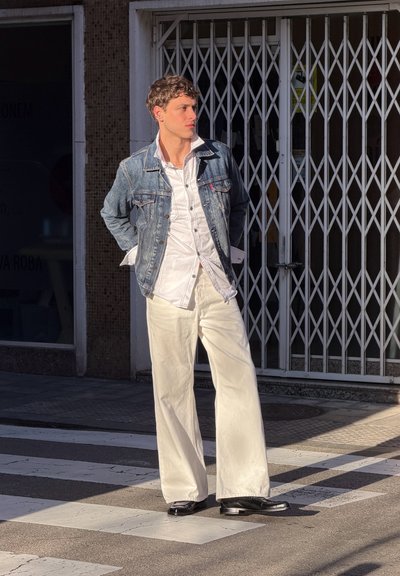 Joven con cabello rizado que lleva una chaqueta vaquera, camisa blanca y pantalones blancos anchos, de pie en un paso de peatones bajo la luz del sol, con las manos detrás de la espalda.