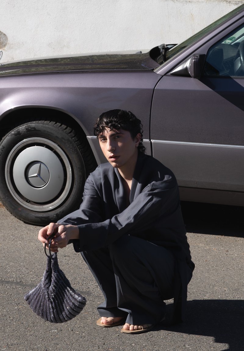 Personne aux cheveux courts et foncés accroupie à côté d'une voiture Mercedes vintage gris foncé, tenant un petit sac texturé noir et argenté dans les deux mains.