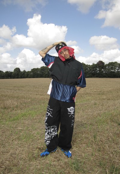 Persona vestida con un chaleco negro sobre una camisa de satén azul, pantalones negros con gráficos blancos, zapatos azules y una bandana roja en un campo.