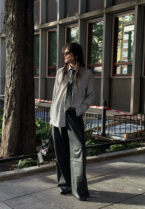 Una mujer con gafas de sol, camisa a rayas, corbata y pantalones de pernera ancha está de pie junto a un árbol, frente a un edificio moderno.