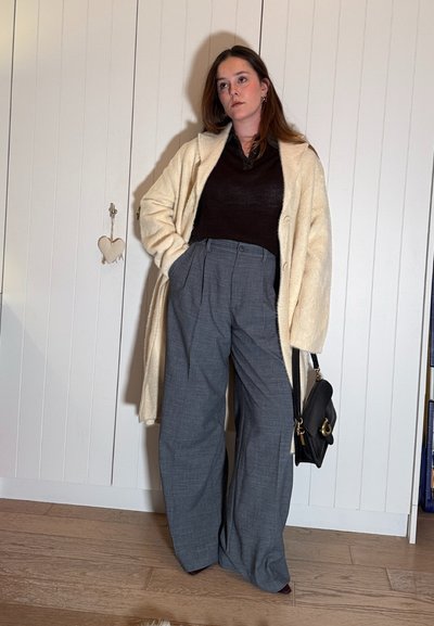 Mujer de pie en interiores que lleva un abrigo color crema, camiseta negra, pantalones anchos grises, sostiene un bolso negro, con las manos en los bolsillos y expresión neutral.