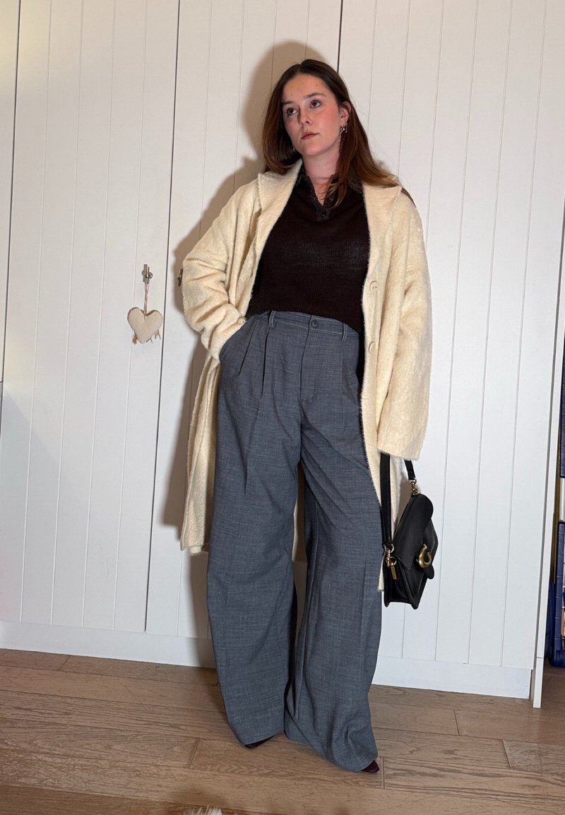 Femme debout à l'intérieur portant un manteau crème, un haut noir, un pantalon à jambes larges gris, tenant un sac à main noir, les mains dans les poches, expression neutre.