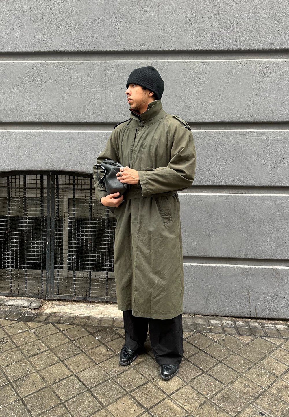 Πράσινο, μακρύ παλτό με ψηλό γιακά και επωμίδες στους ώμους; μαύρο σκούφο; κρατάει μια μαύρη θήκη; φορεμένο με φαρδιά μαύρα παντελόνια και παπούτσια.