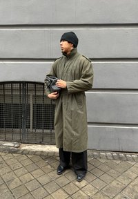 Πράσινο, μακρύ παλτό με ψηλό γιακά και επωμίδες στους ώμους; μαύρο σκούφο; κρατάει μια μαύρη θήκη; φορεμένο με φαρδιά μαύρα παντελόνια και παπούτσια.