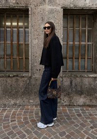 Femme aux cheveux longs et raides portant une veste noire, un jean large et foncé, des baskets blanches, des lunettes de soleil, et tenant un sac à main coloré en perles dans une rue pavée.