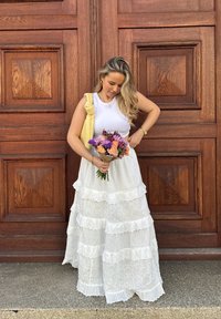 Weißes, gestuftes Maxikleid mit Spitzenakzenten, kombiniert mit einer gelben, gesteppten Tasche. Hält einen Strauß rosa und lila Blumen. Holz Hintergrund.
