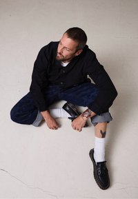 Camisa preta de botões, calças largas de padrão escuro, meias brancas e sapatos de couro pretos com franjas. Tatuagens notáveis no braço.