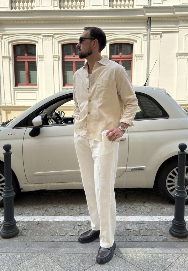 Homme en chemise beige et pantalon blanc tenant une tasse de café se tient à côté d'une petite voiture blanche dans une rue de la ville avec des fenêtres d'immeubles classiques en arrière-plan.