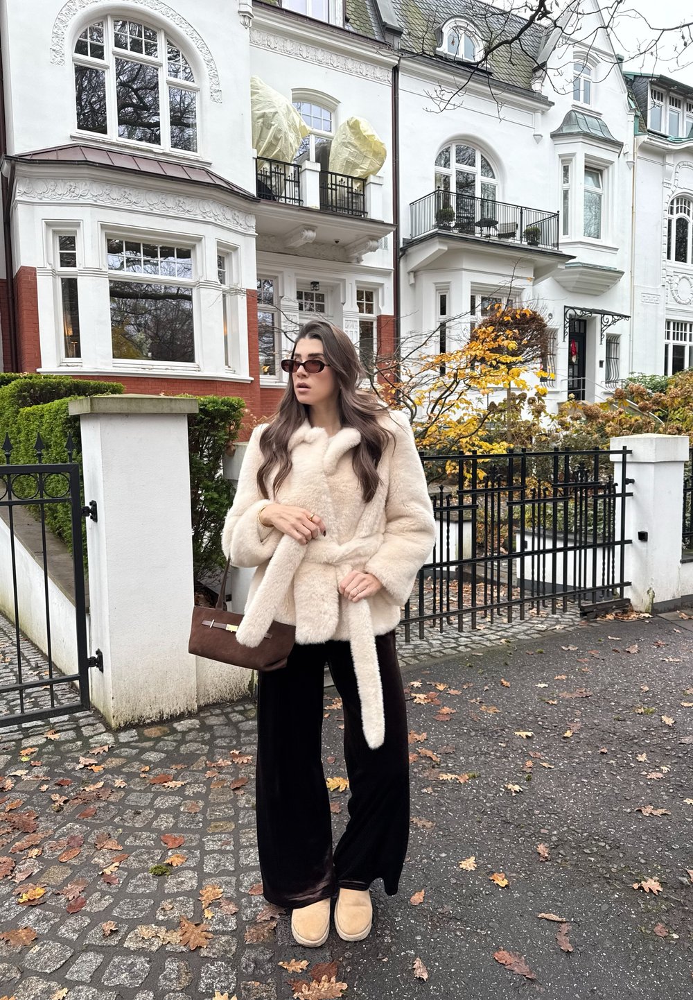 Femme portant un manteau en fourrure beige et un pantalon noir, tenant un sac à main marron, debout sur une rue pavée couverte de feuilles devant des maisons blanches en rangée.