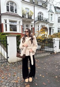 Femme portant un manteau en fourrure beige et un pantalon noir, tenant un sac à main marron, debout sur une rue pavée couverte de feuilles devant des maisons blanches en rangée.